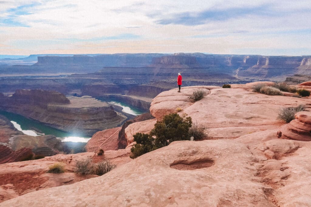 Person in red coat standing at the edge of a cliff with panoramic views of the canyon and river below in Dead Horse Point State Park
