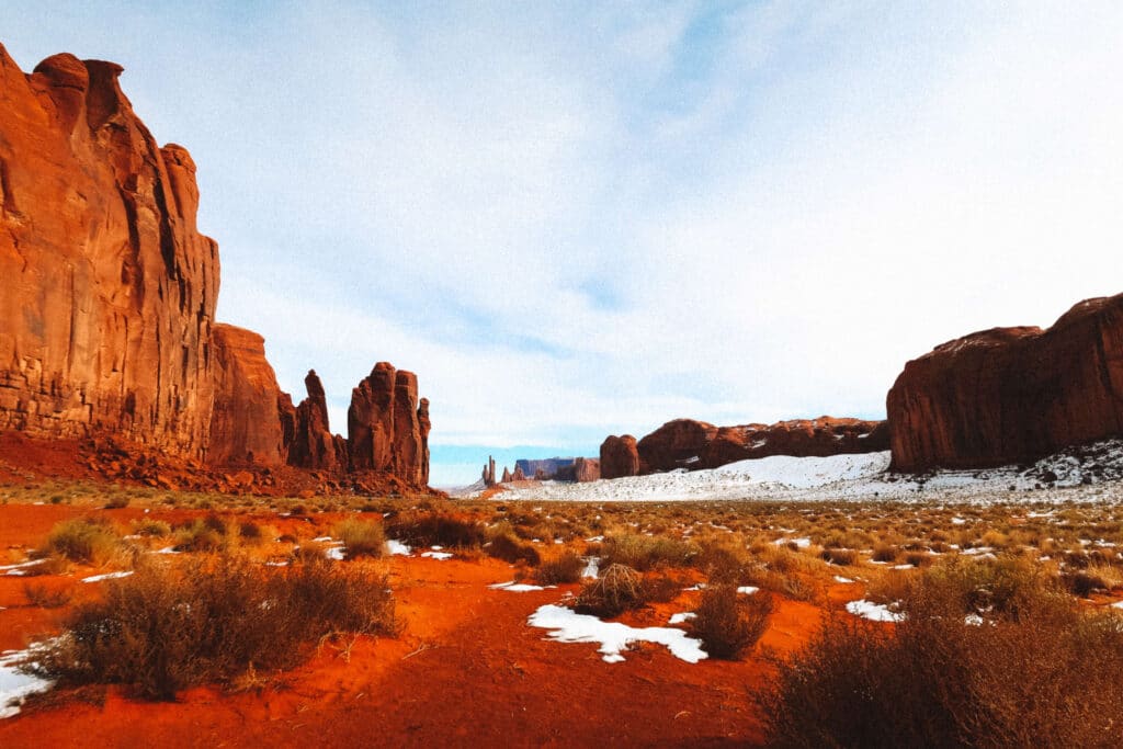 Layered sandstone cliffs and brush with patchy snow and a bright blue sky above.