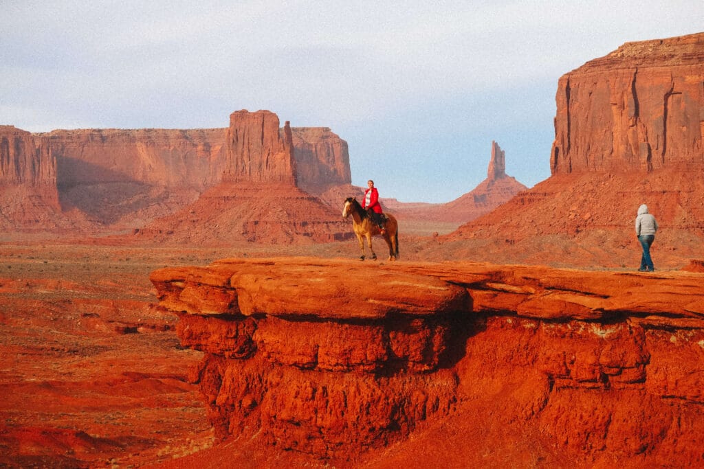 A travelers sitting on a horse standing on a red rock ledge gazing toward distant buttes and mesas on the Monument Valley Loop 17 Mile Drive