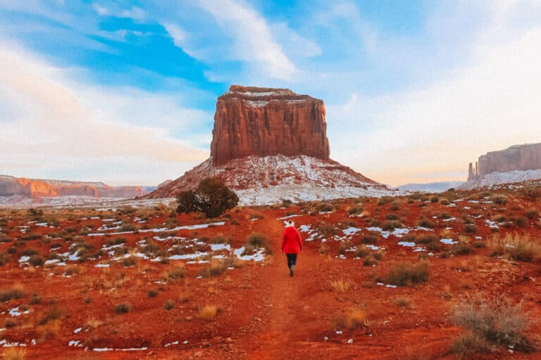A figure in a red parka walking along a snowy desert trail toward a massive butte under a dramatic sky.