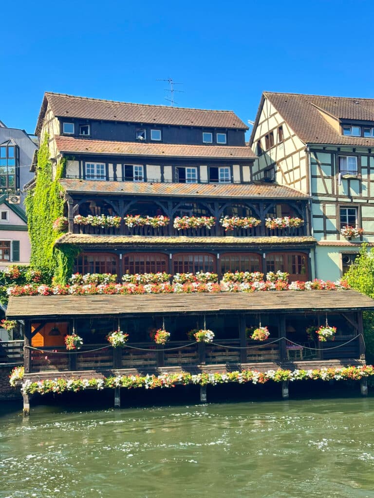 Multi-story building with cascading flowers on balconies overlooking the river in Strasbourg’s historic center.