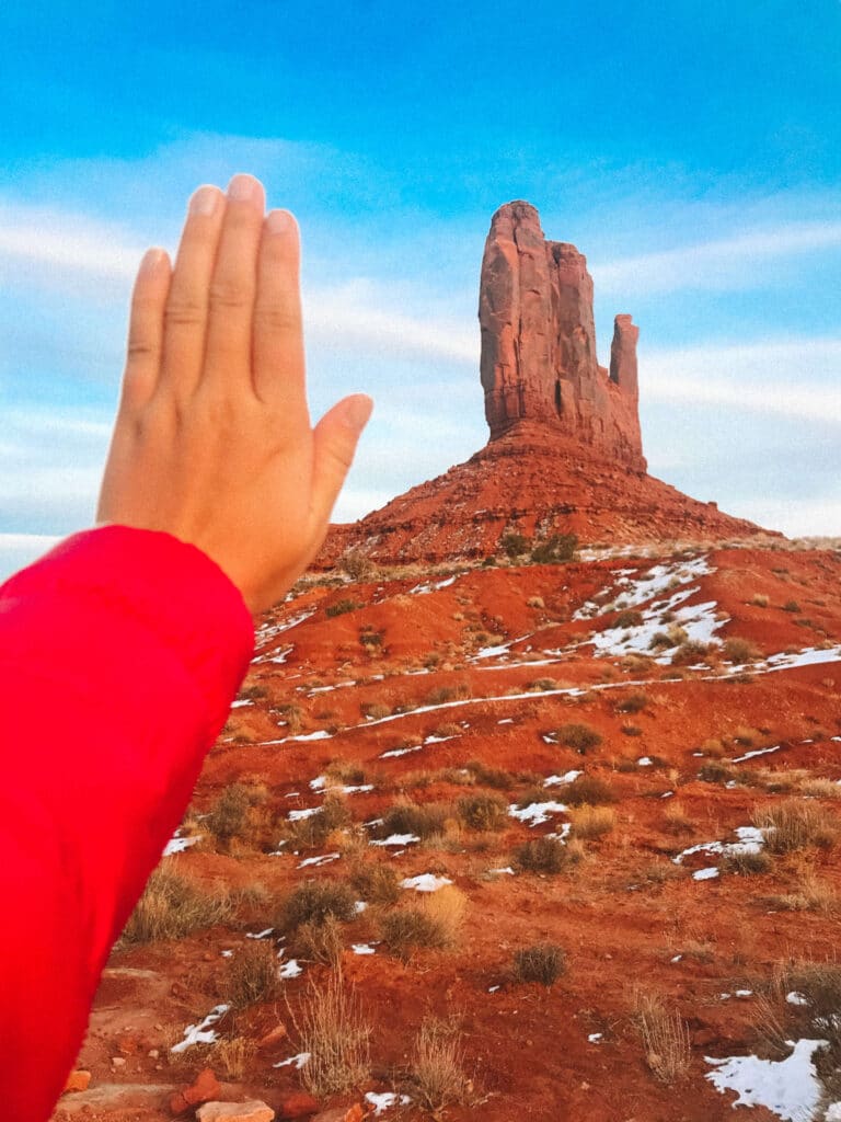 A hand held up in the foreground playfully mimicking the shape of a mitten rock formation, with snow-speckled desert landscape in the background.