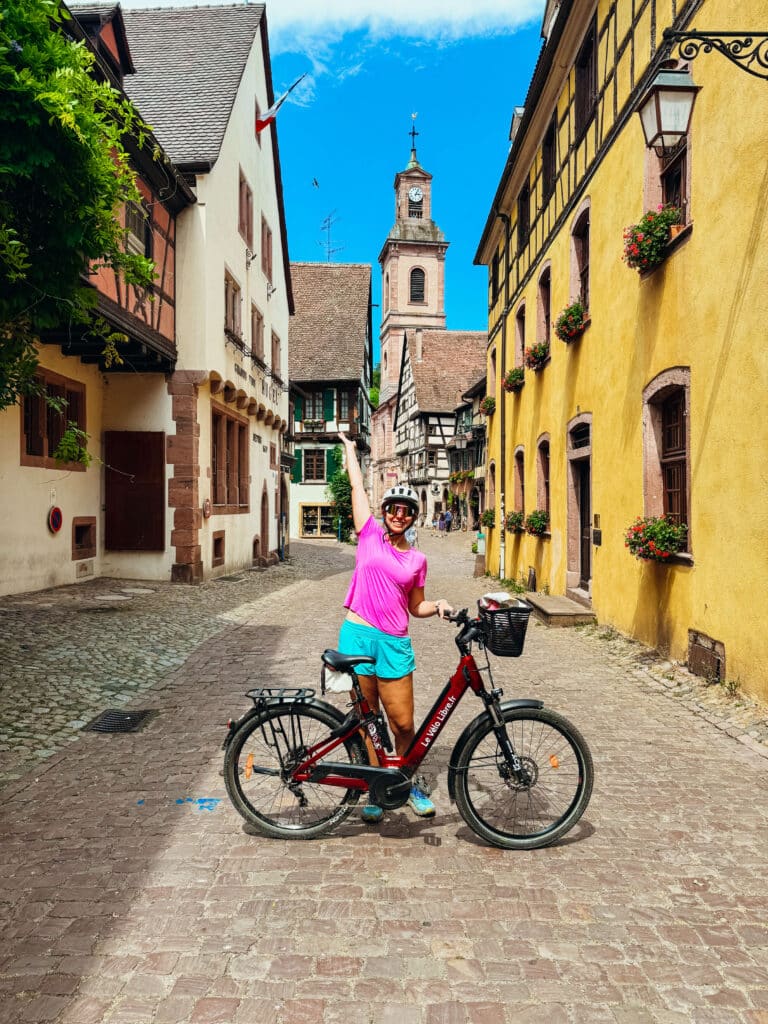 Woman posing on a bike the a colorful Alsace village of Riquewihr with a church tower in the background