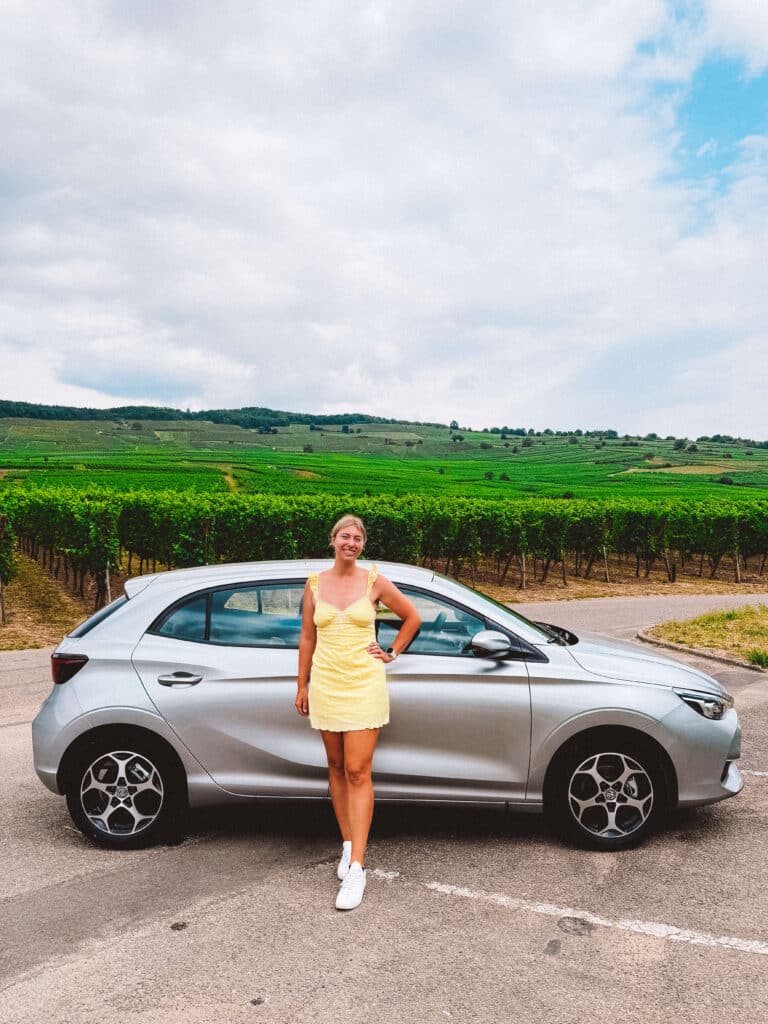 Woman in a yellow dress standing beside a silver rental car parked in front of lush green vineyards in Alsace, France.