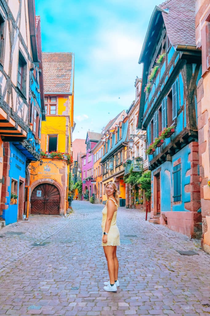 Colorful cobblestone street in Riquewihr, France, with a smiling woman in a pale yellow dress — a peaceful scene that hides what came next.