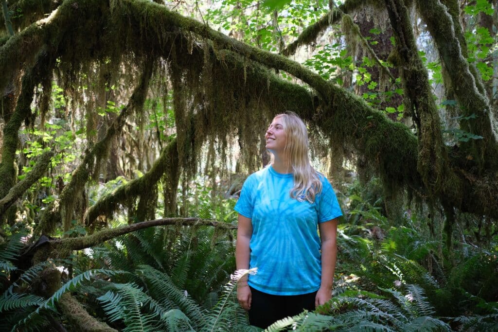 A woman in a blue tie-dye shirt smiles while standing beneath moss-draped tree branches in Hoh Rainforest.