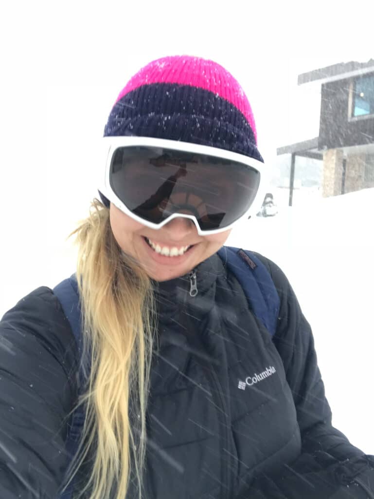 Smiling woman in ski goggles and a pink hat taking a selfie in the snow, with ski lodge buildings in the background.