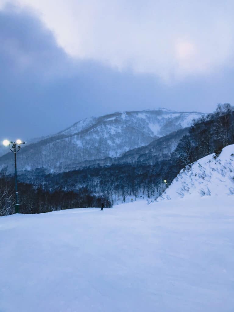 Evening view of a snowy mountain slope under a moody blue sky, with ski lights illuminating the scene.