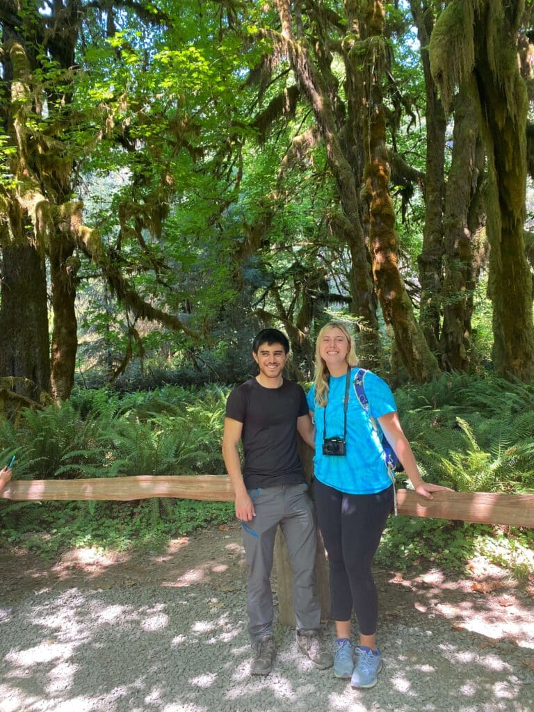 Two hikers smile for the camera while standing on a forest path, surrounded by dense greenery at Hoh Rainforest