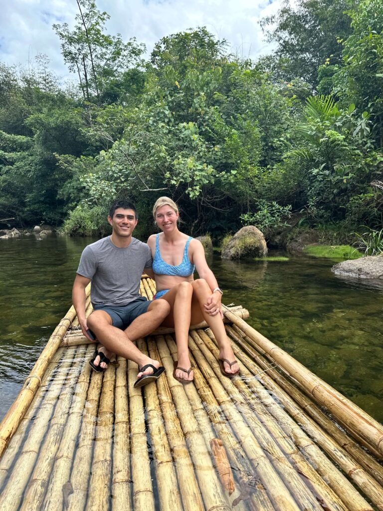 A smiling couple sits at the front of a bamboo raft, surrounded by tranquil river and dense green jungle.