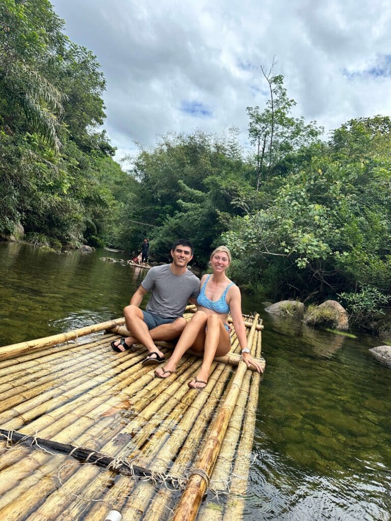 A couple poses on a bamboo raft in the jungle in Thailand, with overhanging trees and soft natural light