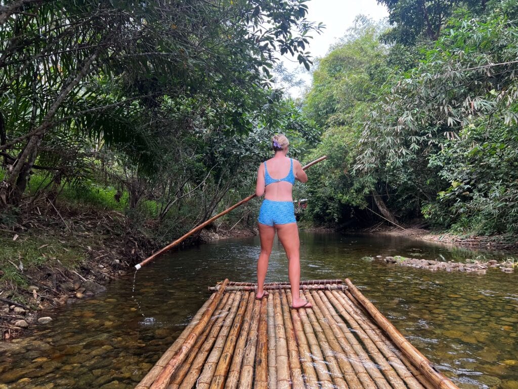 a woman continues rafting downstream, bamboo pole in hand, surrounded by tall trees and clear skies.