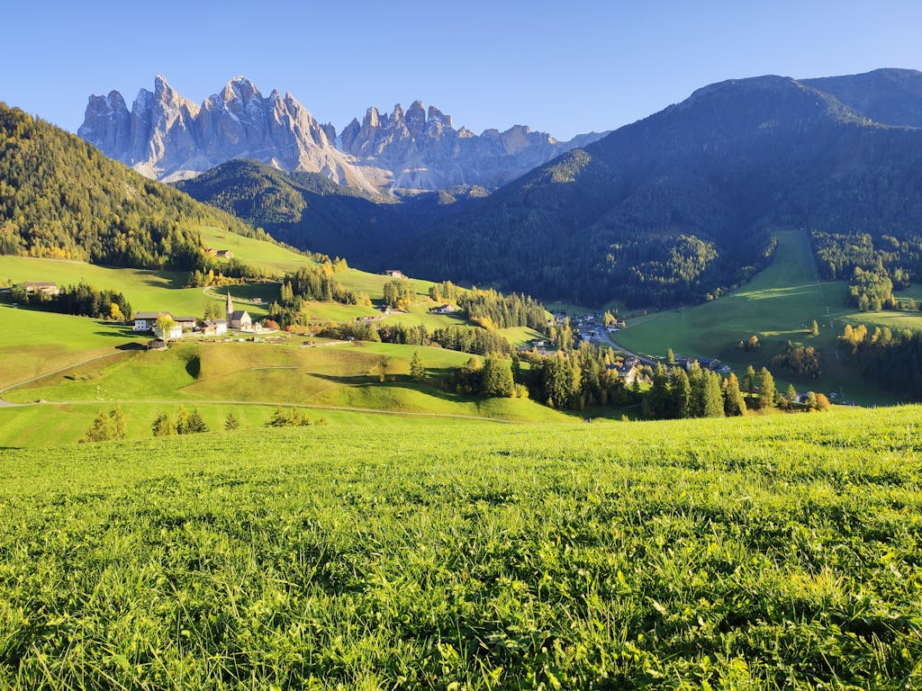 Breathtaking landscape of the Dolomites, lush valleys, and rural houses under a clear blue sky.