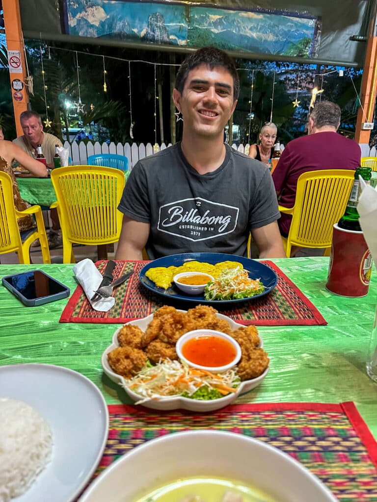Traveler smiling at an outdoor table at Give Me Five Restaurant in Khao Lak, with colorful plates of Thai food.