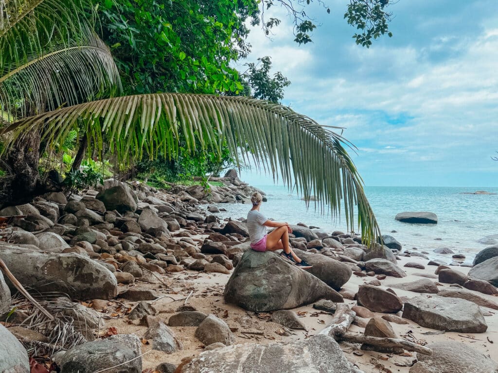Traveler sitting on a large boulder under a leaning palm tree at a rocky beach in Khao Lak–Lam Ru National Park