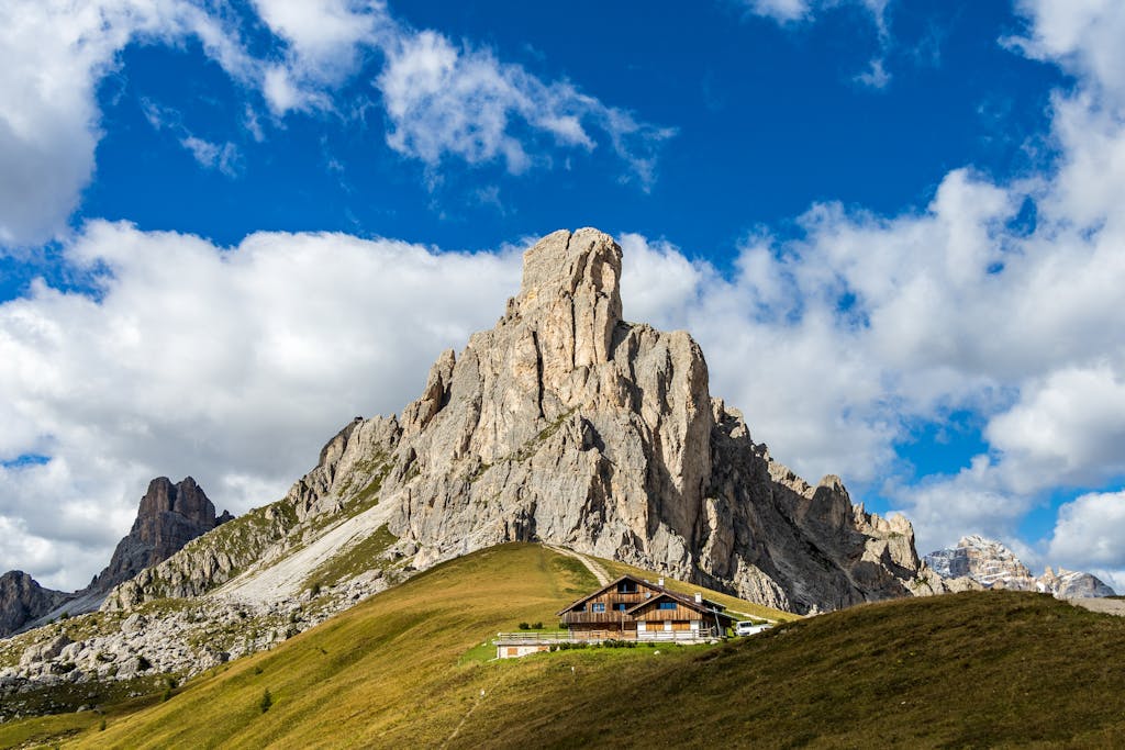 Picturesque scenery of wooden houses placed on grassy hill near rocky Dolomite Mountains against cloudy blue sky in daylight