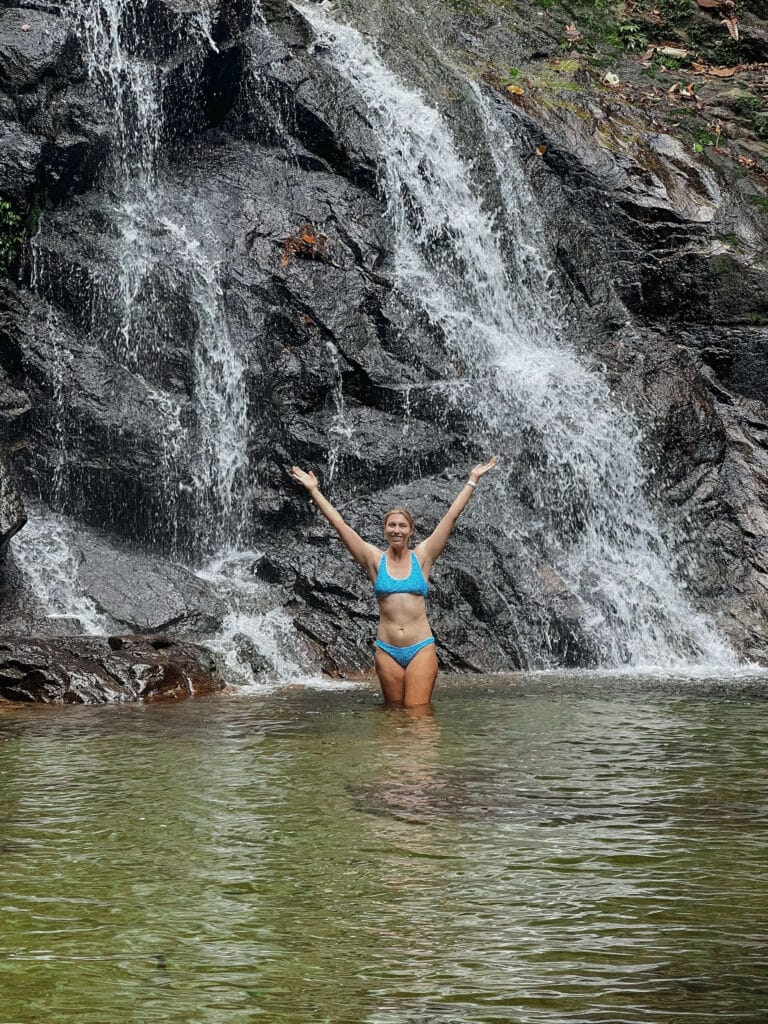 Traveler joyfully raising arms while waist-deep in a waterfall pool, surrounded by rocky cliffs.