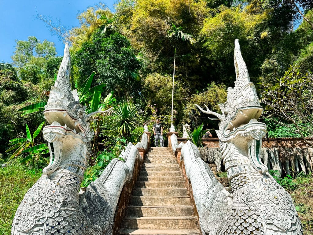 Ornate naga dragon statues lining the stairway to Wat Pha Lat temple, surrounded by jungle.