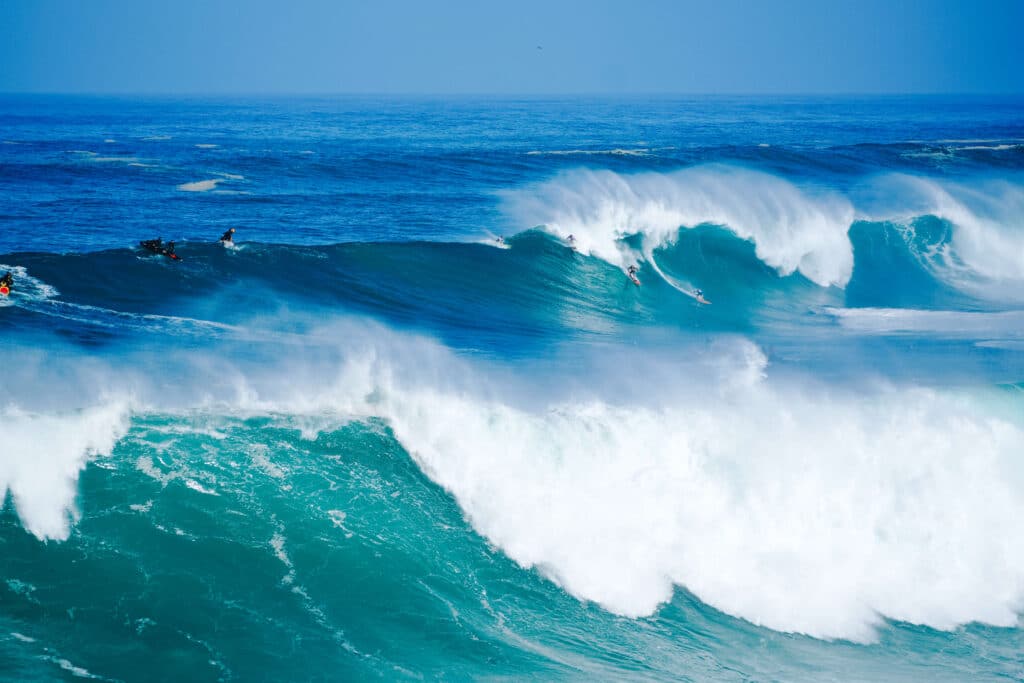 Massive rolling swell at Waimea Bay crashing into the shore under bright blue skies.