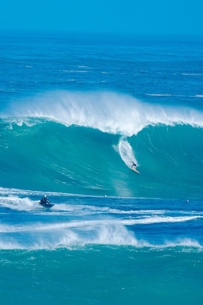 Surfer riding a giant wave with the steep face of turquoise water rising behind them.