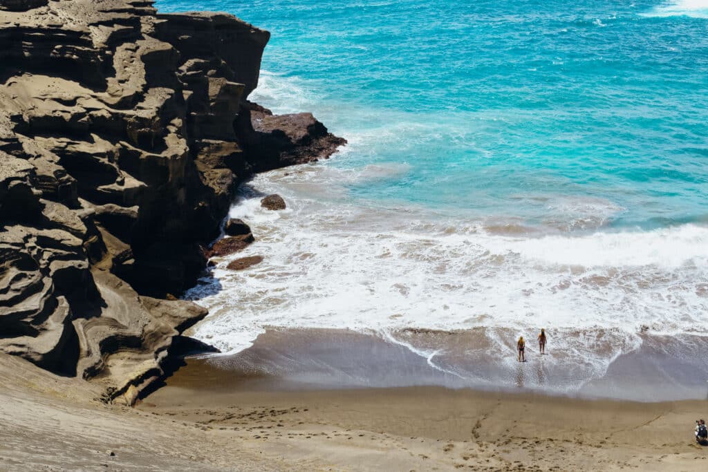 Close-up of sharp lava rock formations beside the shoreline, with waves breaking below against the greenish sand.