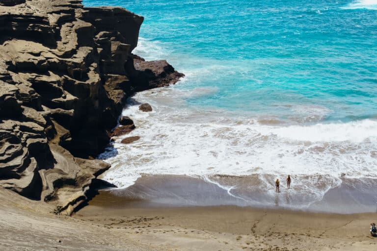 Close-up of sharp lava rock formations beside the shoreline, with waves breaking below against the greenish sand.