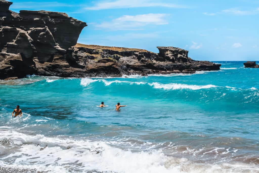 People swim in bright blue water beneath large, rugged lava cliffs at Papakōlea Green Sand Beach.