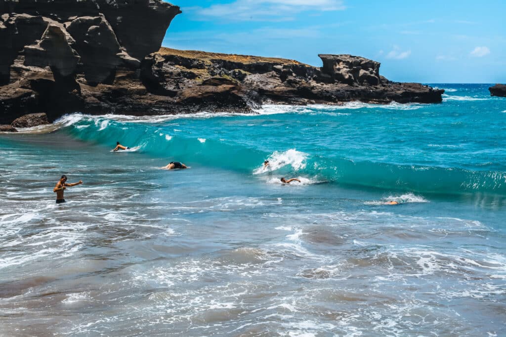 Swimmers bob in choppy turquoise surf as waves break hard against the narrow green-tinted shoreline.