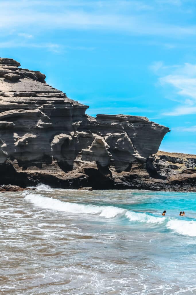 Waves roll onto Papakōlea’s greenish sand beneath a tall lava bluff, with bright blue sky overhead.