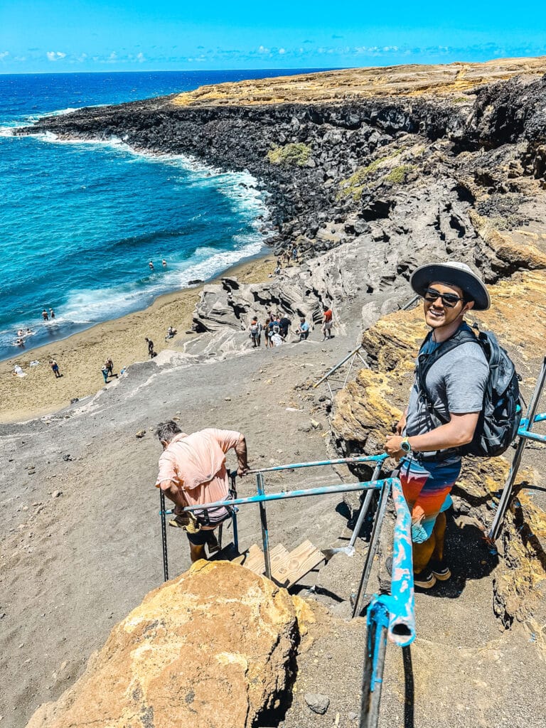 A hiker stands near the edge of the cinder cone overlooking Papakōlea Green Sand Beach, with the turquoise bay and steep ladder stails of the trail visible below.