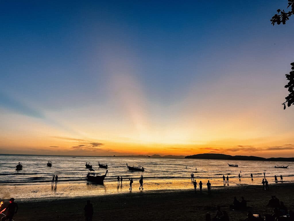 Silhouetted longtail boats anchored on the water at Ao Nang as the sun sets, casting golden and orange light across the horizon.