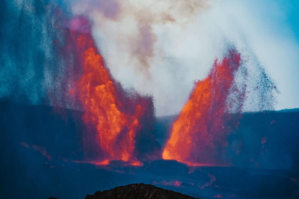 A dramatic nighttime lava fountain glowing orange against dark volcanic cliffs.