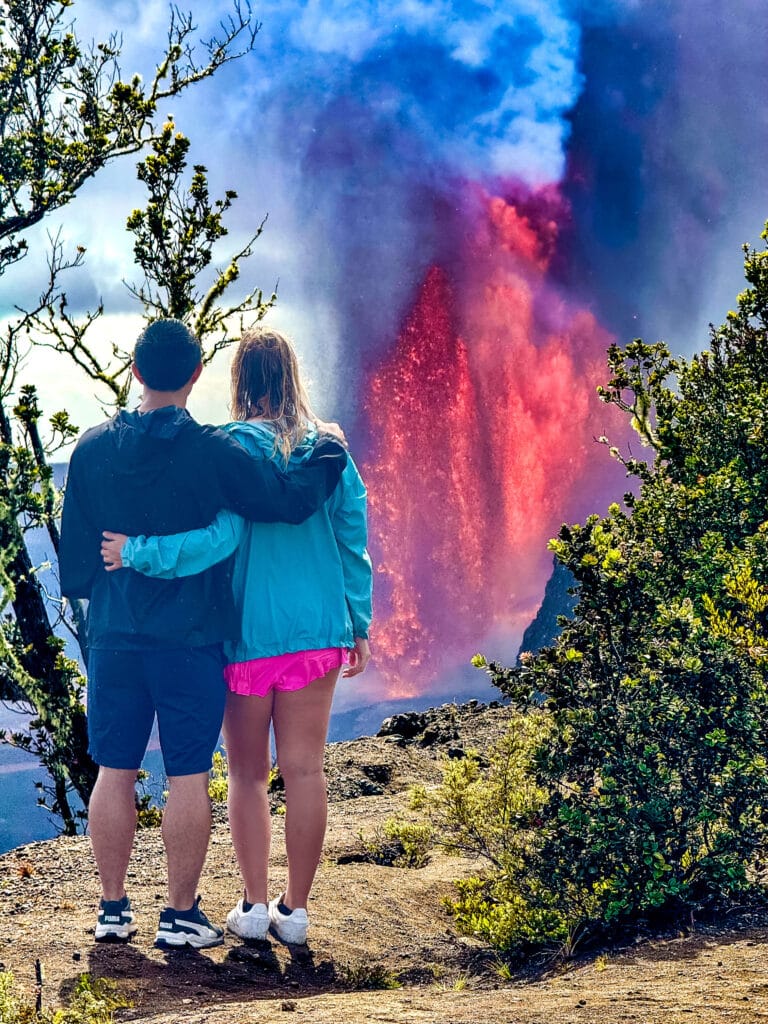 Two people standing at a lookout, watching a tall, bright lava fountain erupting inside the Kīlauea caldera.