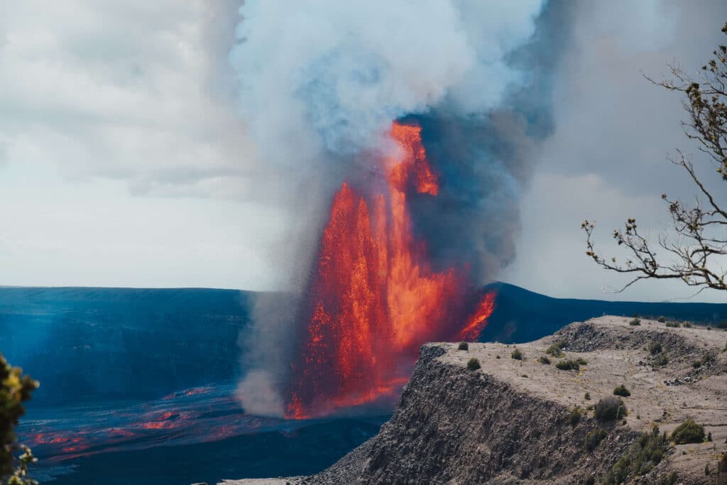 A powerful lava fountain shooting upward as seen from the Kīlauea Overlook, with rugged crater walls behind it.