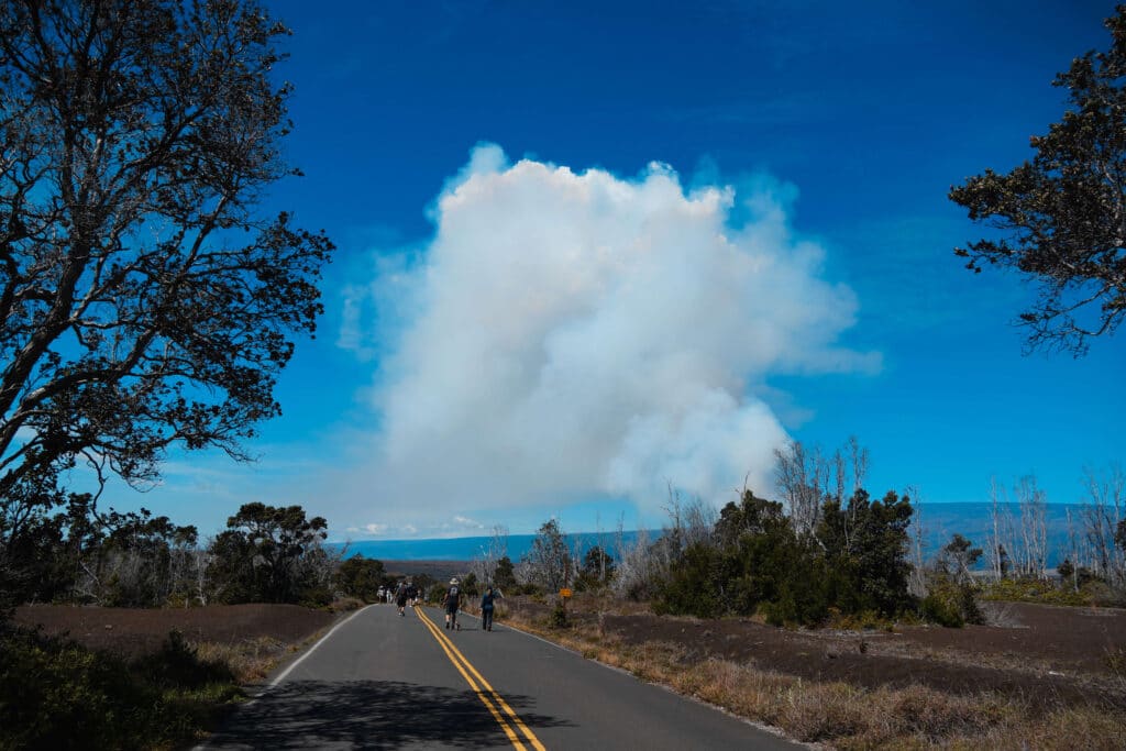 A quiet, closed road leading toward a huge plume of volcanic steam in the distance.