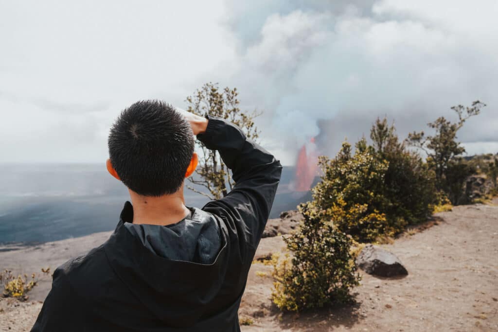 A tourist photographing a plume of volcanic smoke rising from the crater on a cloudy day.