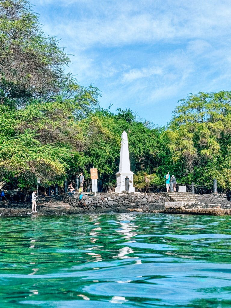 The white Captain Cook Monument standing at the edge of Kealakekua Bay surrounded by greenery.