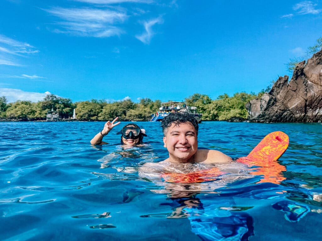 A smiling snorkeler floating in bright blue water near the cliffs of Kealakekua Bay on a sunny morning.
