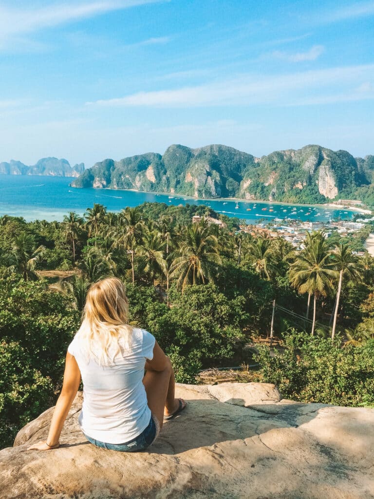 A traveler sitting on a rock at the Phi Phi viewpoint, overlooking turquoise water and green islands.