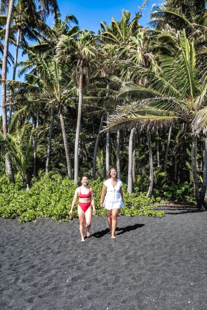 Two women walking along the black sand at Punaluʻu Beach, surrounded by tall palm trees and lush greenery.