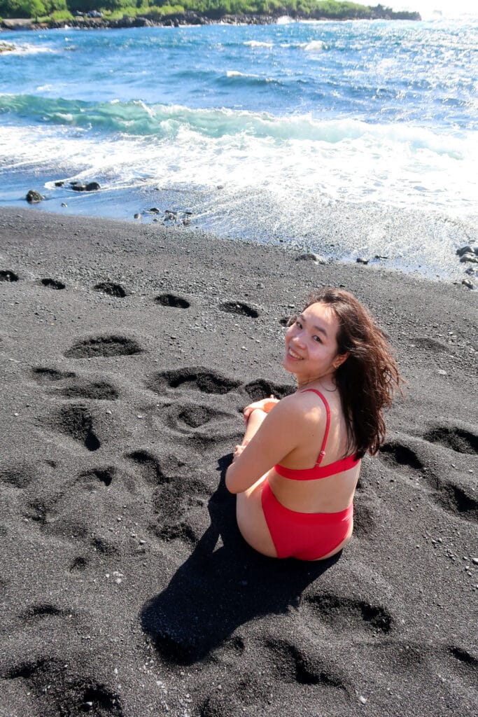 A woman sitting on the black sand at Punaluʻu Beach, framed by footprints and bright blue surf under sunny skies.