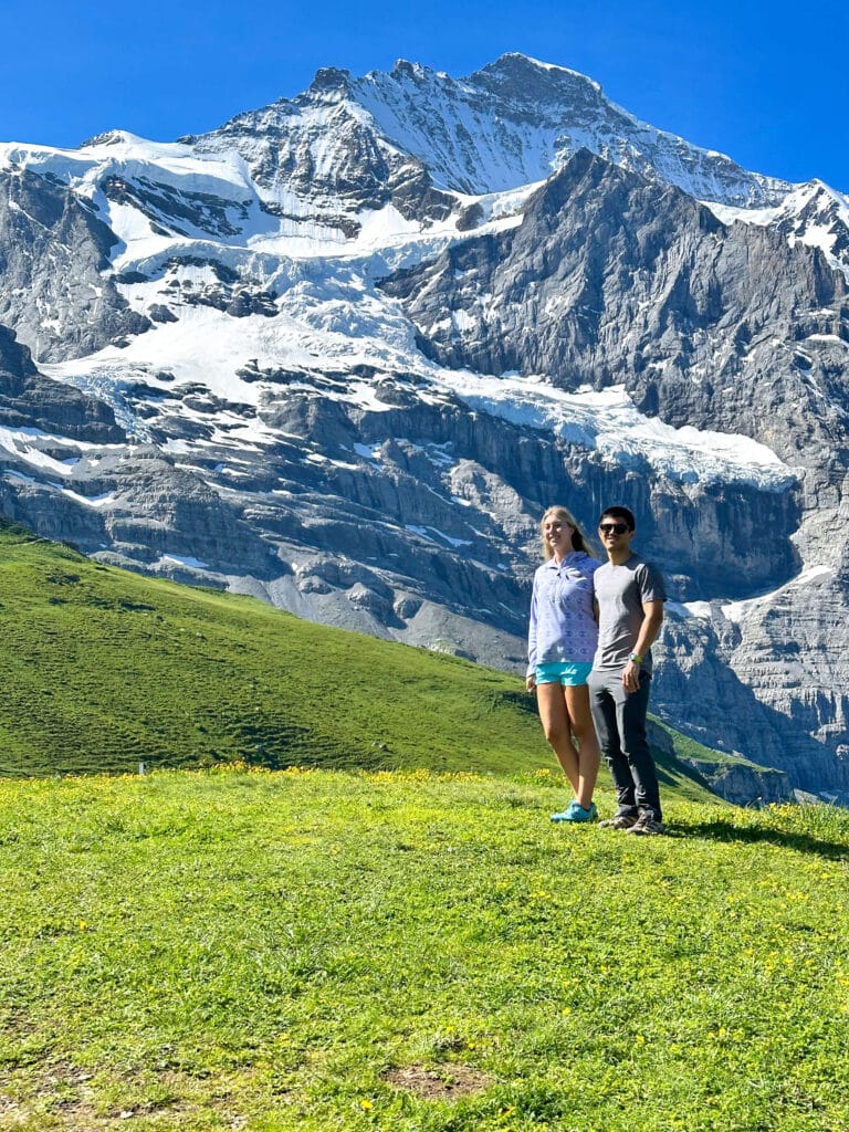 Two travelers standing on an alpine meadow with the Eiger’s North Face towering behind them