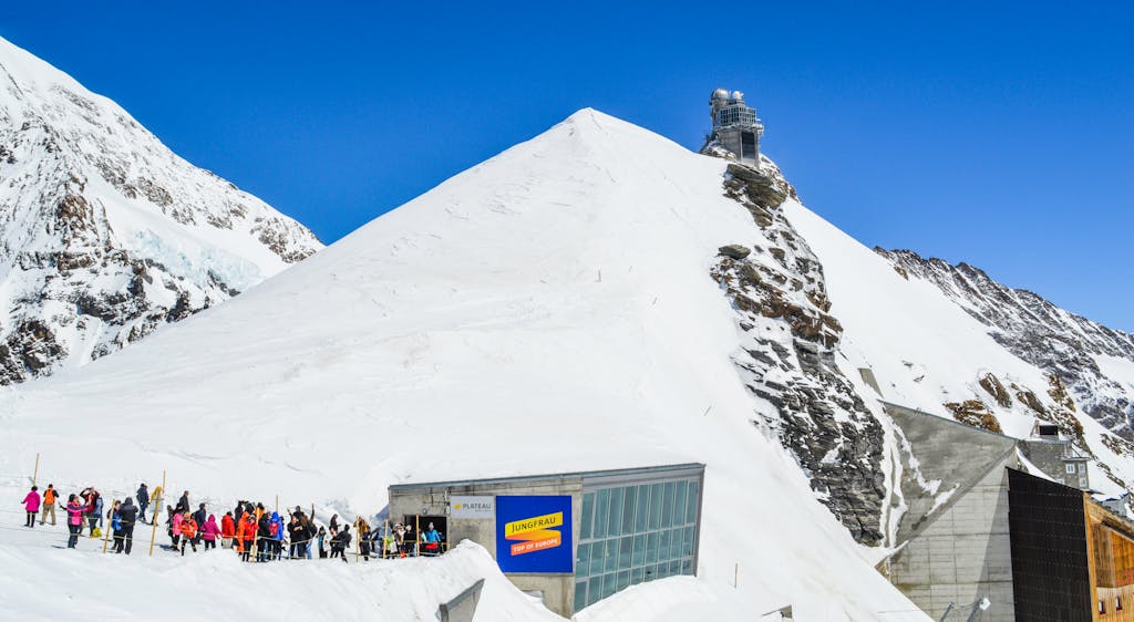 Anonymous group of tourists with skis near old building on snowy mountain under blue sky