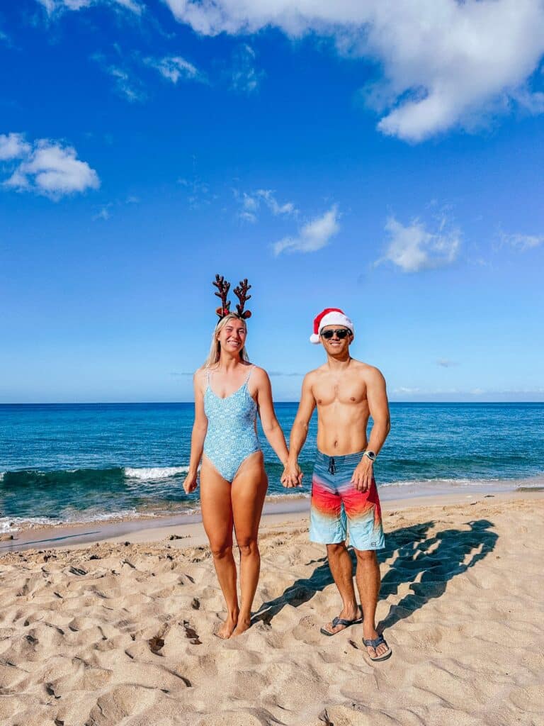 Two people wearing Santa hats and reindeer antlers pose on a sunny Hawaiian beach with palm trees and blue sky in the background.