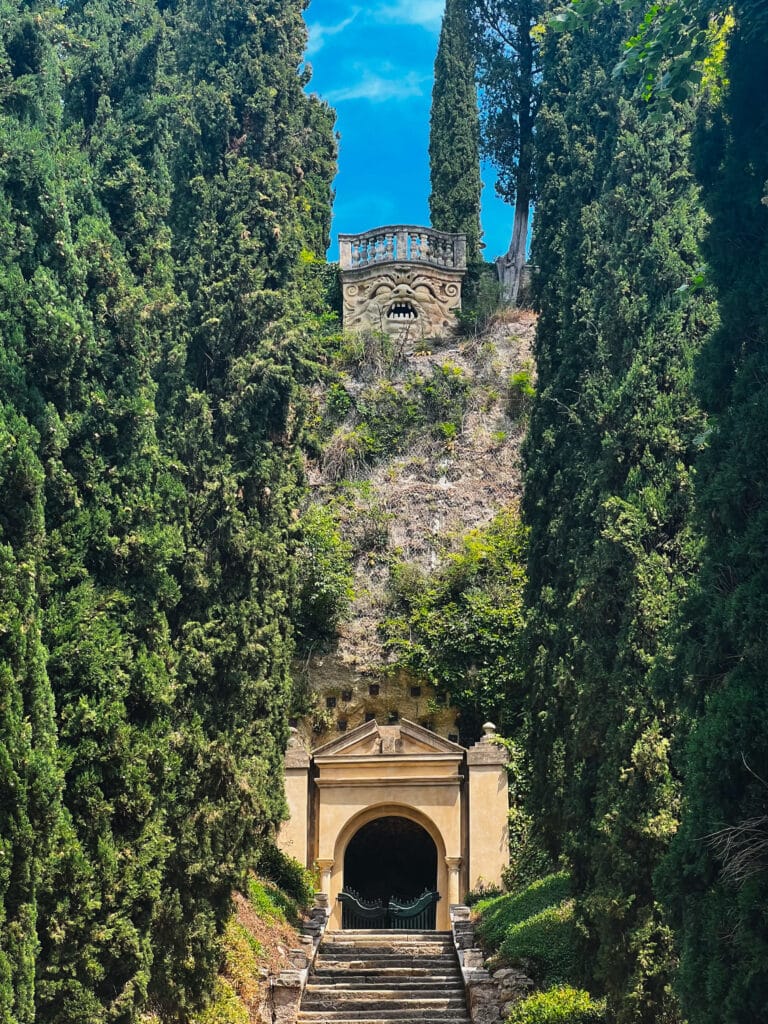The grotesque stone mask carved into the hillside above the grotto at Giardino Giusti, flanked by tall cypress trees and a small tower