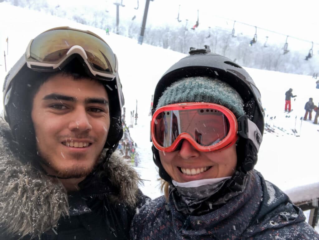 Two bundled-up skiers taking a selfie on a snowy day in Hakuba, Japan, with white ski slopes in the background.