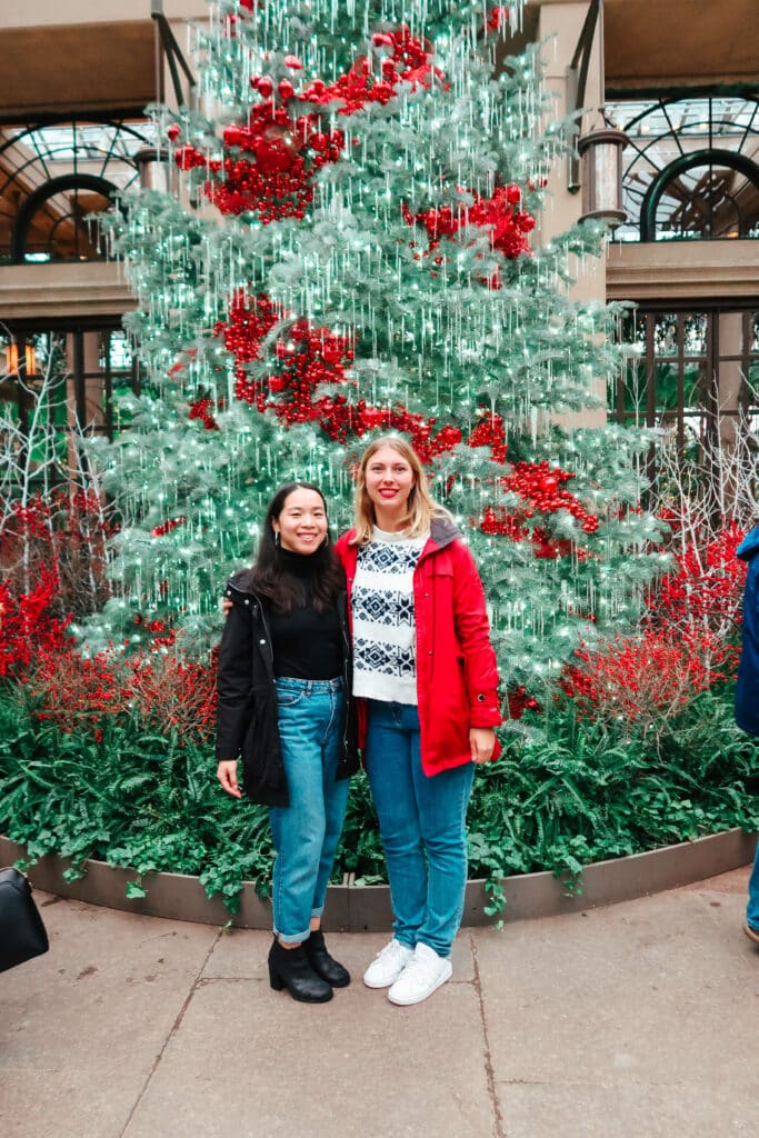 Two friends posing in front of a massive indoor Christmas tree decorated with oversized ornaments at Longwood Gardens.
