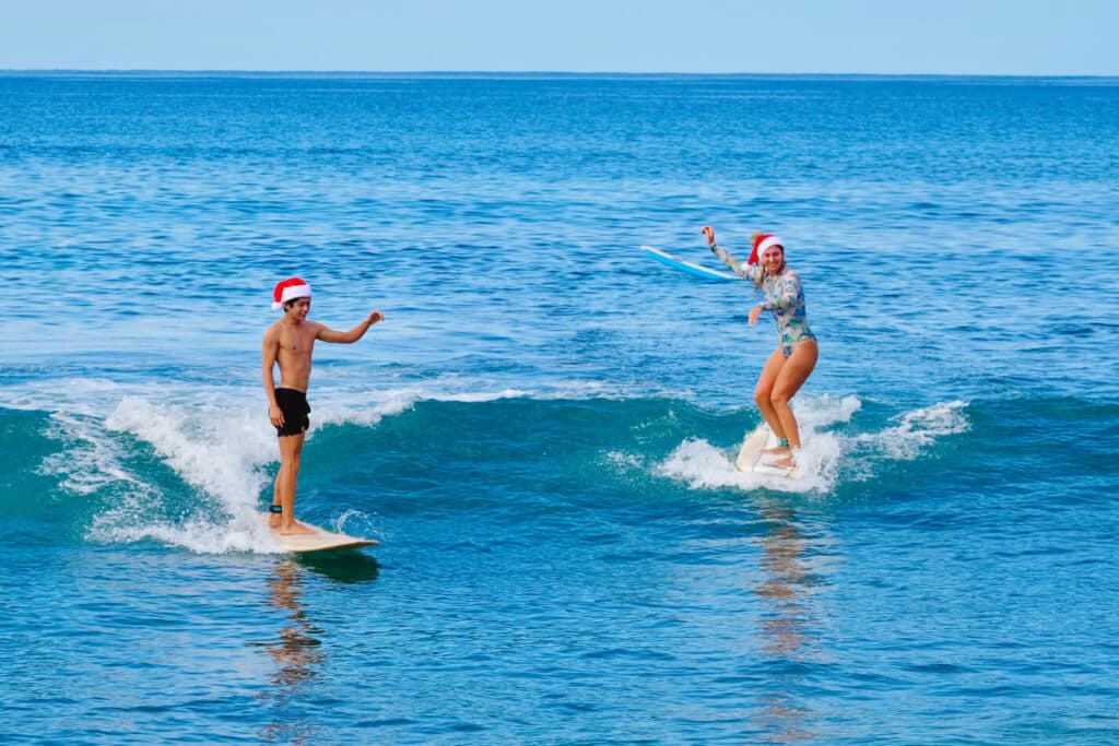 Two surfers in Santa hats ride gentle waves in warm Hawaiian waters, bringing a festive twist to the beach scene.