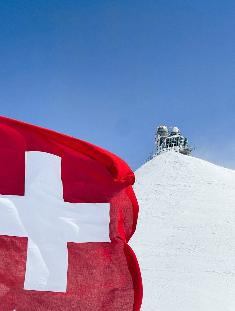 Swiss flag waving in front of the Sphinx Observatory on a snowy peak, under a clear sky.