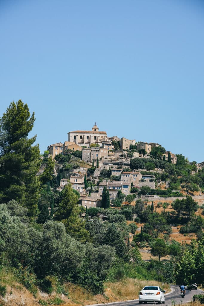 View of Gordes hilltop village in Provence from a roadside lookout, with olive trees and a car on the road below.